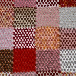 May include: Close-up of a patchwork textile with a variety of colors and textures. The design features squares of pink, red, brown, and white, with open weave and solid sections. The edges have a striped border.