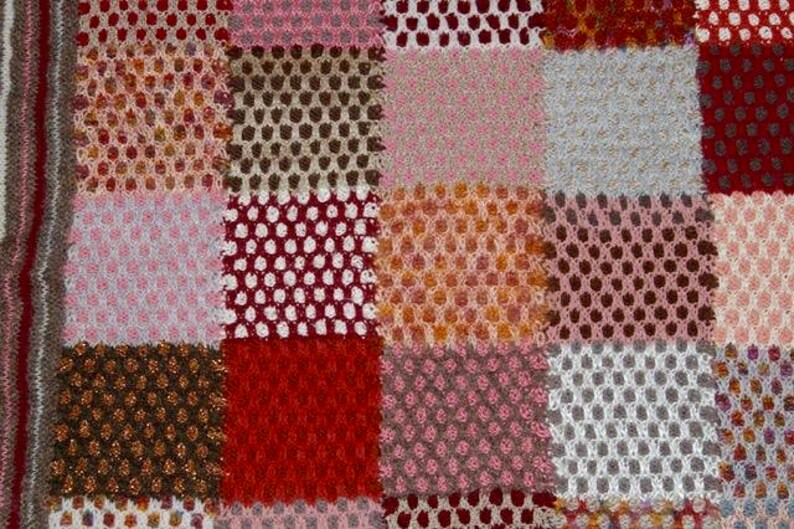 May include: Close-up of a patchwork textile with a variety of colors and textures. The design features squares of pink, red, brown, and white, with open weave and solid sections. The edges have a striped border.