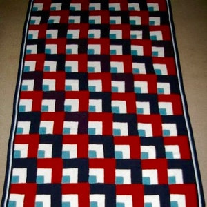 May include: A rectangular, handmade blanket with a geometric pattern. The design features alternating squares of red, navy blue, white, and teal. The blanket is bordered with a navy blue trim. The overall aesthetic is modern and cozy.