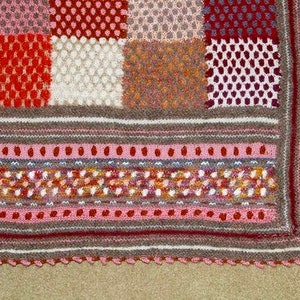 May include: A handmade, multi-colored knitted blanket with a patchwork design. The blanket features squares of various textures and colors, including red, white, and brown. The border has a scalloped edge.
