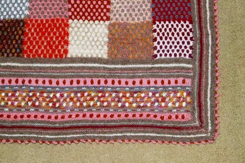 May include: A handmade, multi-colored knitted blanket with a patchwork design. The blanket features squares of various textures and colors, including red, white, and brown. The border has a scalloped edge.