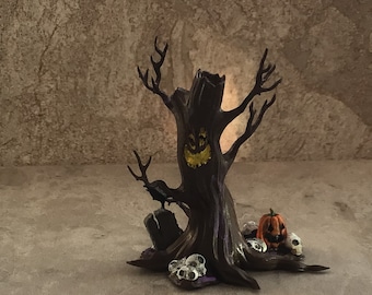 Può includere: Un albero di Halloween inquietante con un viso intagliato, un corvo appollaiato su una lapide e una zucca intagliata alla base. L'albero è circondato da piccoli teschi bianchi.