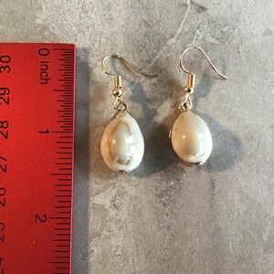 Puede incluir: Un par de pendientes de oro con colgantes de concha blanca. Las conchas son de forma ovalada y tienen un patrón natural. Los pendientes miden aproximadamente 2,5 cm de largo.