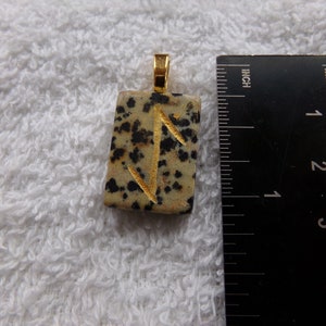 May include: A rectangular pendant with a gold-colored bail. The pendant is a speckled beige stone with black spots and a gold-colored rune symbol. A ruler is next to the pendant, showing inches.