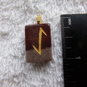 May include: A rectangular pendant with a gold-colored bail. The pendant is a mix of brown and tan with a gold-colored rune symbol. The pendant is approximately 1 inch long.