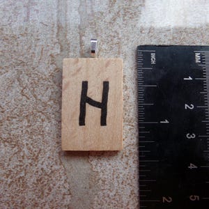 Op de afbeelding: Een houten hanger met de zwarte letter "H". De hanger is rechthoekig en heeft een zilverkleurige beugel. Een liniaal ligt naast de hanger en toont de afmetingen in centimeters.