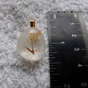 May include: Clear quartz pendant with a gold-colored bail, featuring a gold-colored rune. The pendant is approximately 1 inch in length. The pendant is displayed on a white, textured surface.