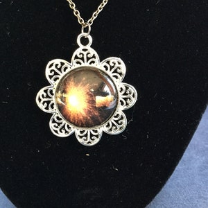 Może przedstawiać: Srebrny naszyjnik z wisiorkiem. Wisiorek ma okrągły, szklany obraz z jasnym żółtym i brązowym wzorem. Wisiorek jest osadzony w ramie w kształcie kwiatu ze skomplikowanymi detalami filigranowymi, zawieszony na delikatnym łańcuszku.