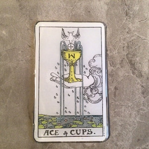 May include: Tarot card featuring the Ace of Cups. The card depicts a golden chalice held by a hand, with a dove above. The card has a white border and the text "ACE of CUPS" at the bottom.
