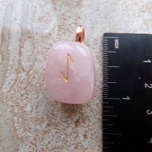 May include: A pink rose quartz pendant with a gold-colored rune symbol. The pendant has a copper-colored loop for a chain. A ruler shows measurements in inches and millimeters.