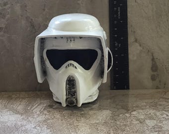 Op de afbeelding: Een witte helm met een zwart vizier en een luchtinlaat aan de voorkant. De helm heeft een afgeronde bovenkant en zijflappen. Een liniaal staat naast de helm en toont metingen in centimeters.