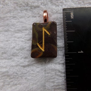 May include: A brown and green pendant with a gold rune symbol, approximately 1 inch in size, on a white towel.