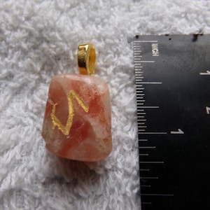 May include: A rectangular pendant with a gold-colored bail. The pendant is a translucent, light-colored stone with red veining and gold-colored accents. A ruler is next to the pendant, showing measurements in inches.