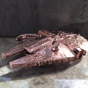 May include: A detailed model of the Millennium Falcon starship, painted in a metallic copper finish. The model is made of plastic and has a realistic design.