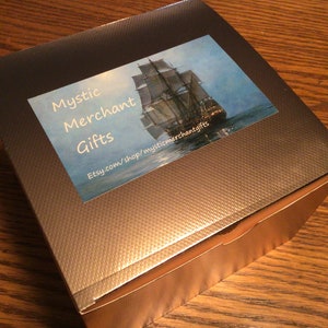 May include: A square gift box with a textured, dark gray lid and a lighter gray base. A sticker on the lid features a painting of a tall ship at sea, with the words "Mystic Merchant Gifts" and a website address.
