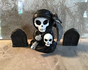May include: A black and white skeletal figure wearing a black hooded robe, holding a skull and a scythe. The figure is sitting in front of two black tombstone decorations with the word "RIP" on one of them.
