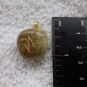 May include: A green stone pendant with a gold-colored bail. The stone has a gold-colored rune symbol on its surface. The pendant is approximately 1 inch in length.