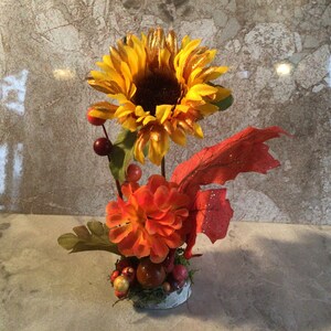 May include: A decorative floral arrangement featuring a large yellow sunflower with a dark brown center, complemented by an orange dahlia and red berries. The arrangement includes green leaves and a red leaf with glitter accents, all set in a small, white-painted container.