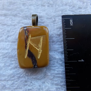 May include: A rectangular pendant with a gold-colored rune symbol on a yellow-brown stone. The pendant has a bronze-colored bail. A ruler shows the pendant is approximately 1 inch in length.