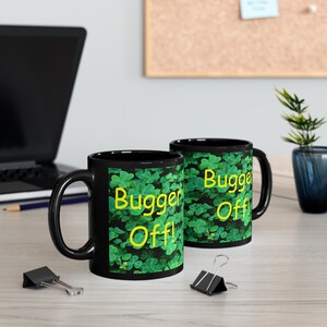 Può includere: Due tazze in ceramica nera con la scritta "Bugger Off!" in giallo. Le tazze presentano un motivo a trifogli verdi. Le tazze sono su una superficie di legno chiaro, con un laptop e forniture per ufficio sullo sfondo.
