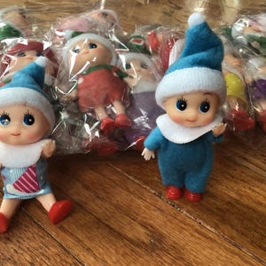 May include: Two small elf dolls with blue hats and outfits, white collars, and red shoes are displayed on a wooden surface. Other elf dolls are visible in the background, some in plastic packaging.