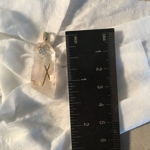 Könnte beinhalten: Ein Anhänger aus klarem Quarzkristall mit einem goldenen Runensymbol. Der Anhänger ist etwa 3,8 cm hoch und hängt an einer goldenen Kette.