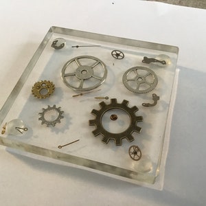 Pode incluir: Um bloco quadrado transparente de resina que contém várias engrenagens e componentes de relógios. As engrenagens são em tons de ouro, prata e bronze, criando uma estética steampunk. O bloco tem cerca de 10 cm de lado.