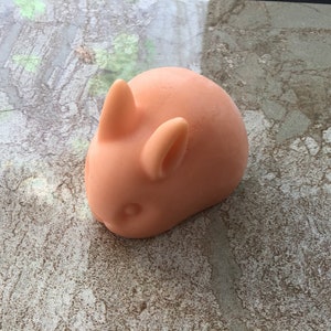 May include: A peach-coloured bunny-shaped object with prominent ears and a rounded body. The toy is smooth and appears to be made of a soft material. The bunny is resting on a textured, mottled surface.