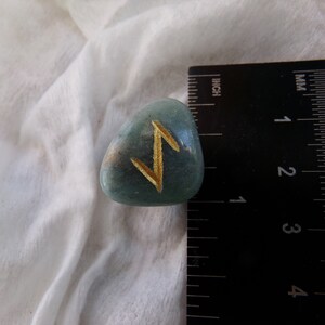 May include: A polished, teal-colored rune stone with a gold-colored symbol. The stone is approximately 1 inch in length and is set against a white fabric background. The rune is a stylized zigzag.