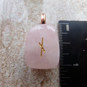 Op de afbeelding: Een gepolijste, lichtroze rozenkwarts hanger met een goudkleurig rune symbool. De hanger heeft een koperkleurige beugel. Een liniaal toont dat de hanger ongeveer 2,5 cm lang is.