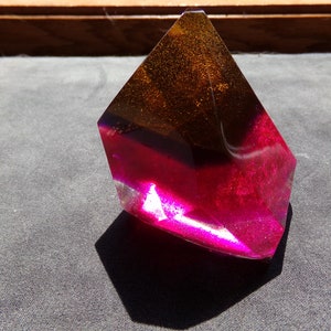 Puede incluir: Una escultura abstracta facetada con un degradado de color. La parte superior es de color ámbar oscuro, que pasa a un magenta vibrante en la base. La escultura está hecha de un material translúcido.