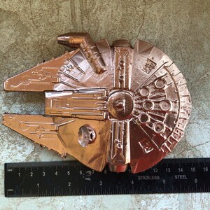 May include: A copper-coloured Millennium Falcon model with detailed surface features. The model is approximately 15 cm long, with a ruler placed next to it for scale. The ruler is black with white markings and the text "STAINLESS STEEL".