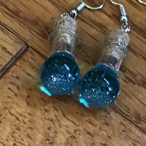 May include: A pair of blue glass bottle earrings with cork stoppers and silver hooks. The bottles are filled with blue glitter.
