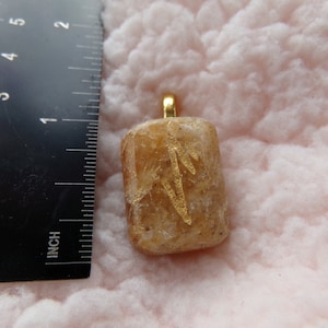 May include: A gold pendant with a rectangular, light brown stone featuring a gold design. The stone is approximately 1 inch in length.