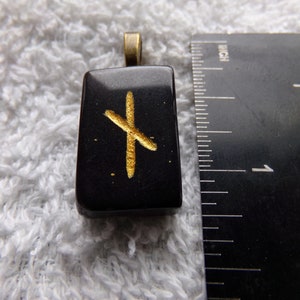 Op de afbeelding: Een rechthoekige zwarte hanger met een goudkleurig rune-symbool. De hanger heeft een bronskleurige beugel om aan een ketting te bevestigen. De hanger is ongeveer 2,5 cm lang.