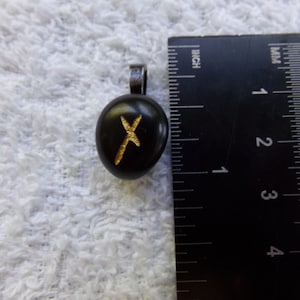 Könnte beinhalten: Schwarzer, ovaler Anhänger mit einem goldfarbenen Runensymbol. Er hat eine kleine Metallschlaufe für eine Kette. Der Anhänger ist etwa 2,5 cm lang.