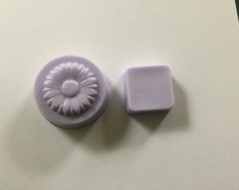 May include: Two lavender-coloured soaps. One is round with a daisy design, the other is a small square. The soaps are on a white surface.