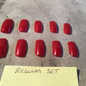Puede incluir: Un conjunto de diez uñas postizas cuadradas de color rojo. Las uñas están dispuestas en dos filas de cinco. El texto "REGULAR SET" está escrito en una nota adhesiva amarilla debajo de las uñas.