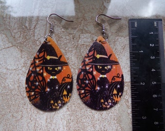 Pode incluir: Par de brincos de Halloween em forma de gota. Cada brinco apresenta um gato preto usando um chapéu de bruxa, em um fundo laranja com teias de aranha e abóboras. Os brincos têm ganchos de orelha prateados.