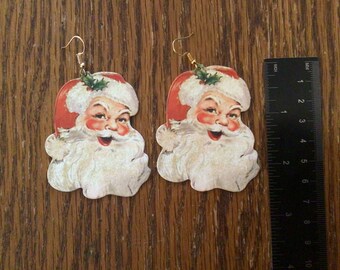 May include: Two Christmas earrings shaped like Santa Claus. Each earring shows Santa's face with a red hat, white beard, and rosy cheeks. The earrings are lightweight and have gold-coloured ear wires. The ruler shows inches.