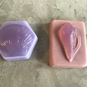May include: Two handmade soap bars with a seashell design. The soap on the left is a light purple color and the soap on the right is a light pink color.