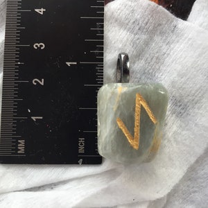 May include: A light green stone pendant with a gold-colored rune symbol. The pendant is approximately 1 inch in length and has a silver-colored bail for attaching to a chain. The stone has a slightly translucent quality.
