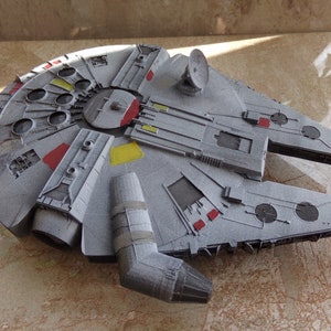 May include: A detailed grey model of the Millennium Falcon starship, with red and yellow highlights. The model shows intricate paneling, a circular radar dish, and various protrusions, capturing the iconic design from the Star Wars franchise.