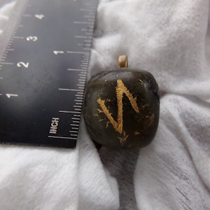 May include: A dark green, square pendant with a gold-colored symbol. The pendant has a small gold loop at the top. A ruler with inch measurements is in the background.