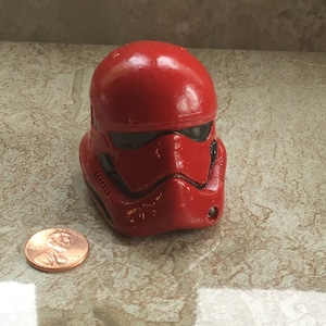 May include: A red Stormtrooper helmet replica with black detailing. The helmet is made of a hard material and has a glossy finish. A penny is next to the helmet for size comparison. The helmet is a collectible item.