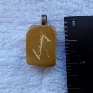 May include: Mustard-colored rectangular pendant with a gold-colored symbol. The pendant has a bronze-colored bail. A ruler is next to the pendant, showing measurements in inches.