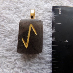May include: A black stone pendant with a gold-colored rune symbol. The pendant is about 1 inch in length and has a gold-colored bail.