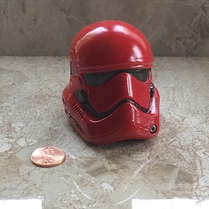 May include: A red helmet, similar to a Star Wars Sith Trooper, with black detailing around the eyes and mouth. A penny is placed beside the helmet for scale. The item is likely a collectible or toy.