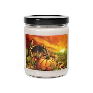 Puede incluir: Un tarro de vela blanco con tapa negra. El tarro presenta una colorida ilustración de una cornucopia rebosante de calabazas, manzanas y otros productos de la cosecha, sobre un fondo de puesta de sol.