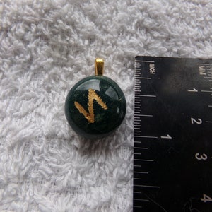 May include: A round, dark green pendant with a gold-colored bail. The pendant features a gold-colored rune. A ruler is next to the pendant, showing measurements in inches.